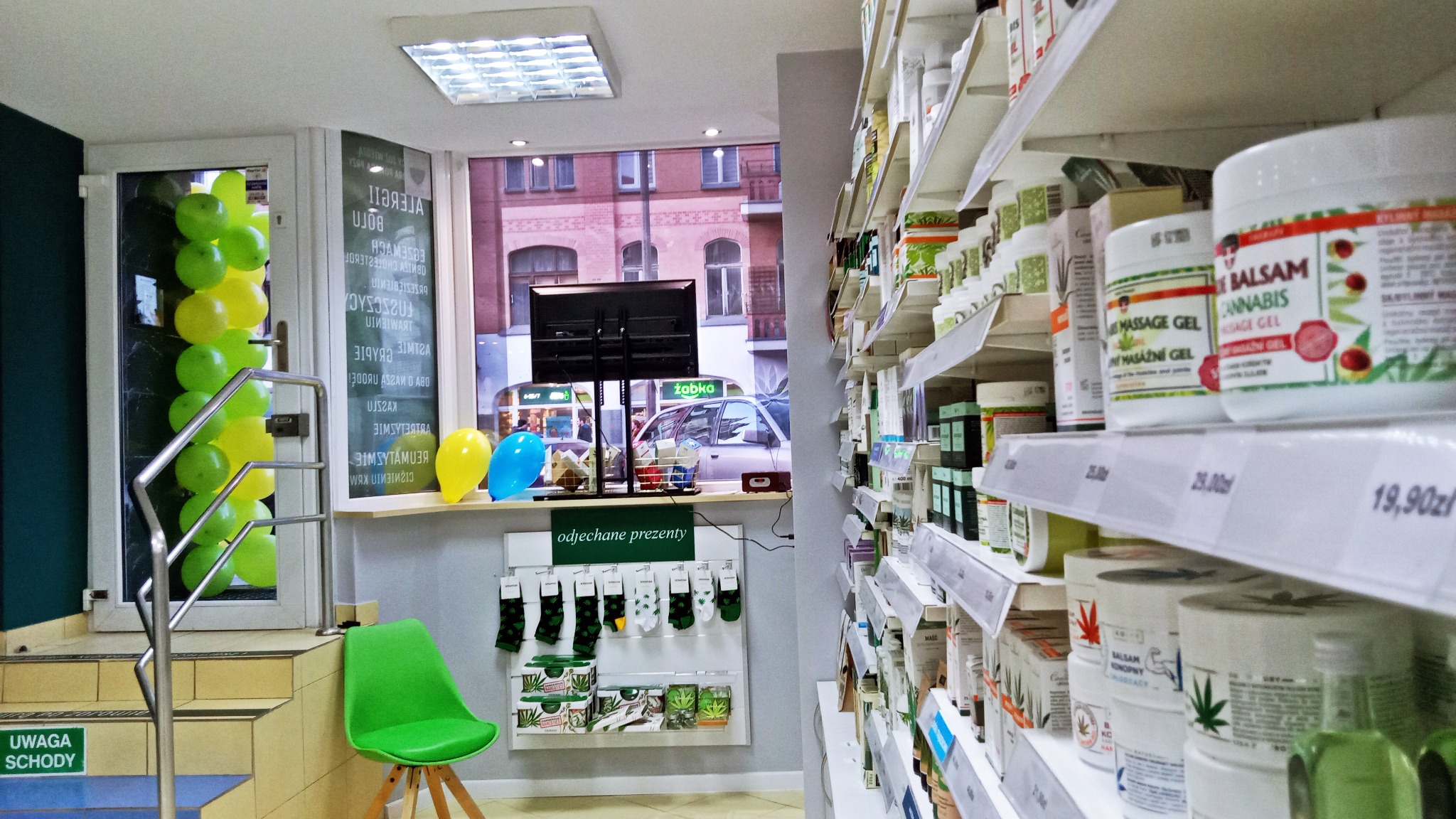 Wnętrze sklepu z produktami naturalnymi: półki wypełnione słoikami i pudełkami z balsamami konopnymi i żelami do masażu, dekoracje z balonów, prezentowe skarpetki z motywem konopi, widok na ulicę...