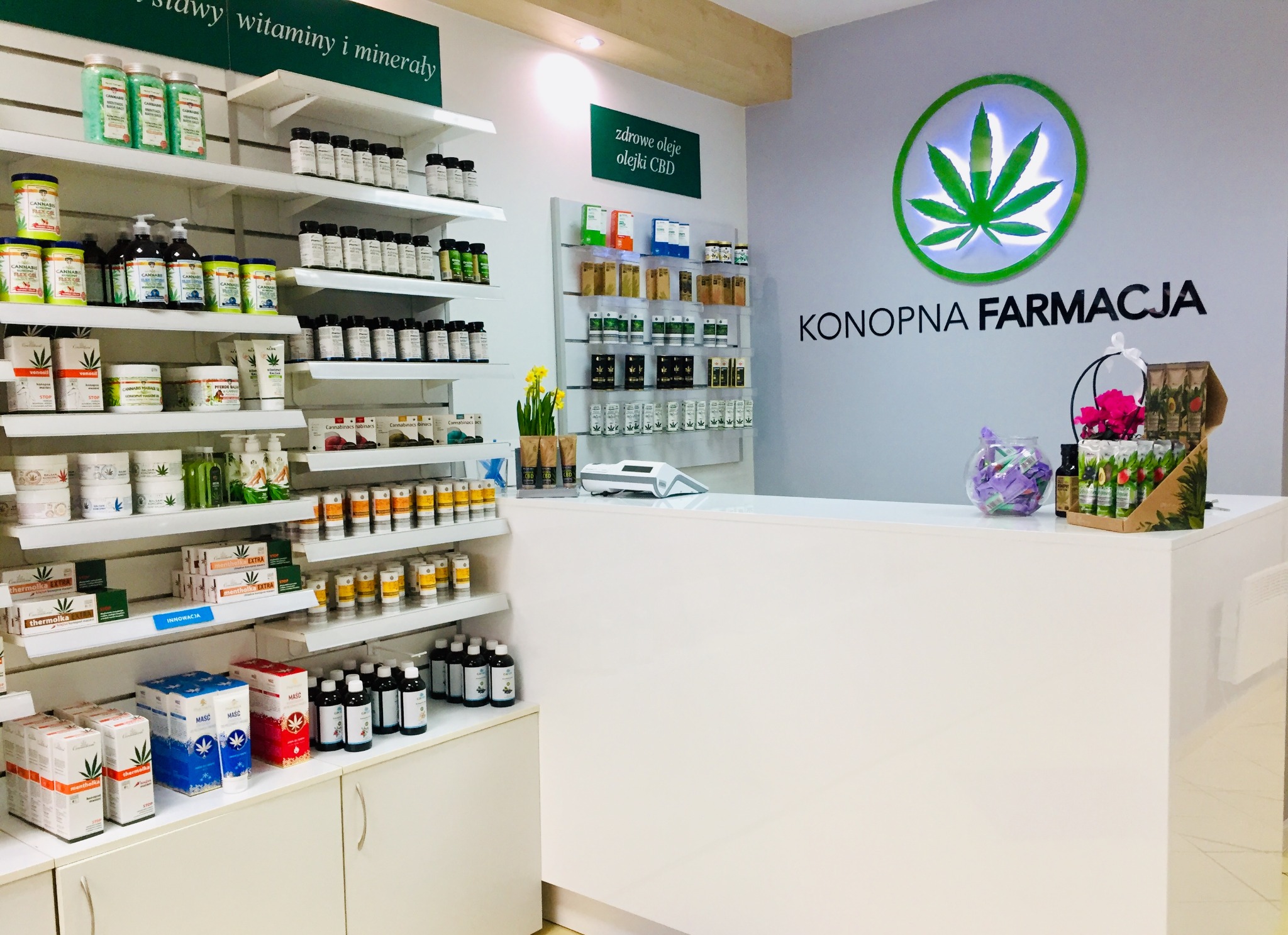 Wnętrze sklepu Konopna Farmacja z półkami pełnymi produktów konopnych, w tym olejków CBD, kosmetyków i suplementów diety, z logo firmy z liściem konopi na ścianie nad białą ladą.