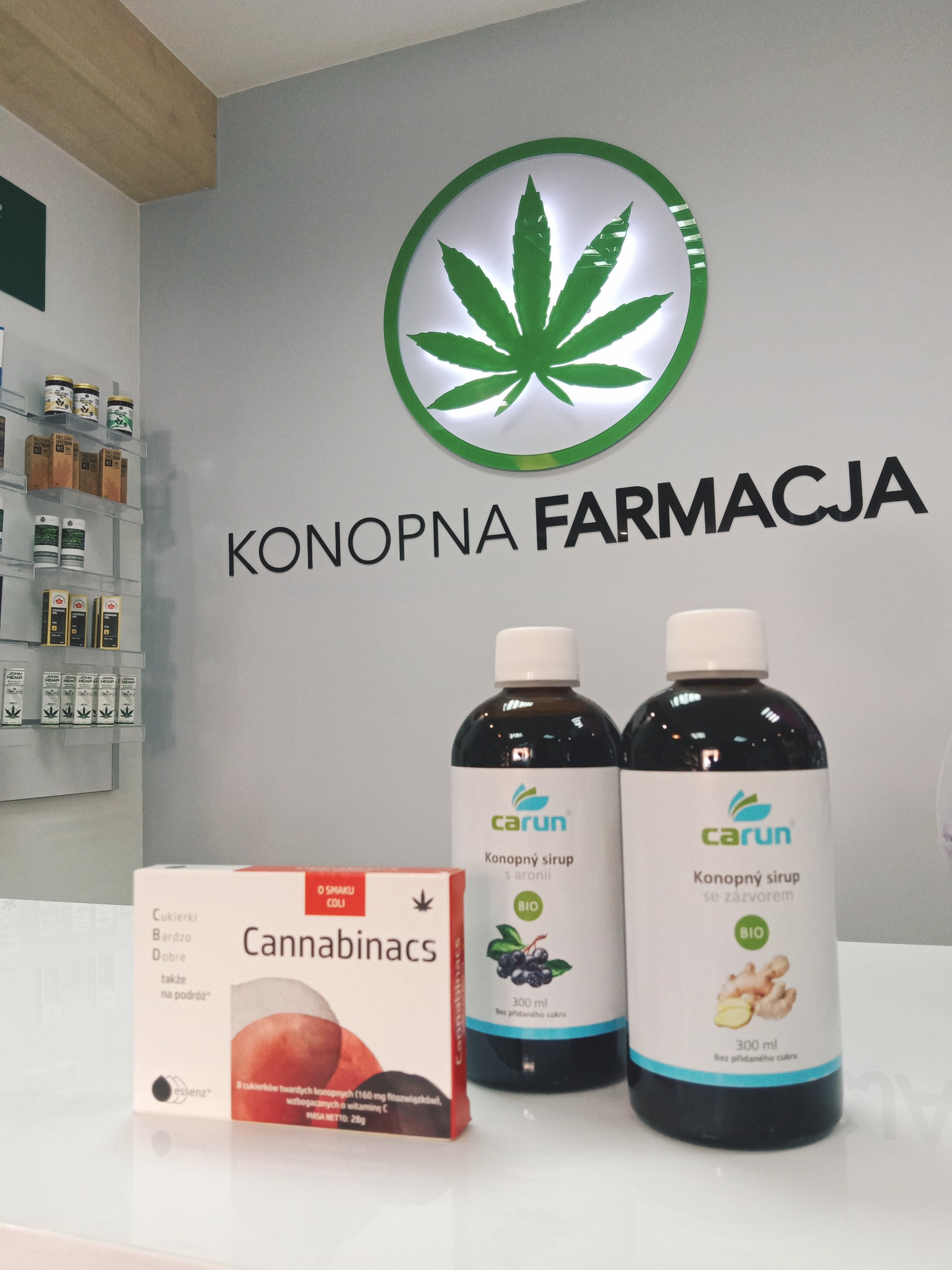 Półka z produktami Konopna Farmacja: syropy konopne Carun z aronią i imbirem oraz cukierki Cannabinacs o smaku coli na białym blacie, w tle logo z liściem konopi.