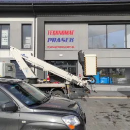 Ciężarówka z podnośnikiem koszowym na tle budynku z dużym logo firmy Technomat Prasek, widok z perspektywy samochodu osobowego.