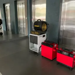 Profesjonalny sprzęt do osuszania: czerwone i białe osuszacze powietrza oraz wentylator Stanley na tle nowoczesnego wnętrza z windami.