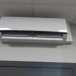 Biały klimatyzator ścienny z wyświetlaczem LED wskazującym temperaturę 30 stopni Celsjusza, zamontowany nad ciemną framugą drzwiową w pomieszczeniu o jasnych ścianach i sufitach kasetonowych.