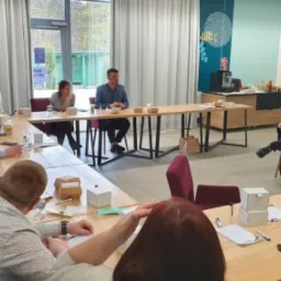 Grupa osób siedząca przy stole w kształcie litery U podczas spotkania lub szkolenia w jasnym pomieszczeniu. Jeden mężczyzna siedzi na krześle na końcu stołu.