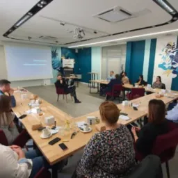 Sala konferencyjna podczas prezentacji: grupa osób siedzi przy stole w kształcie litery U, prelegentka siedzi na krześle przed ekranem projekcyjnym z logotypem firmy.