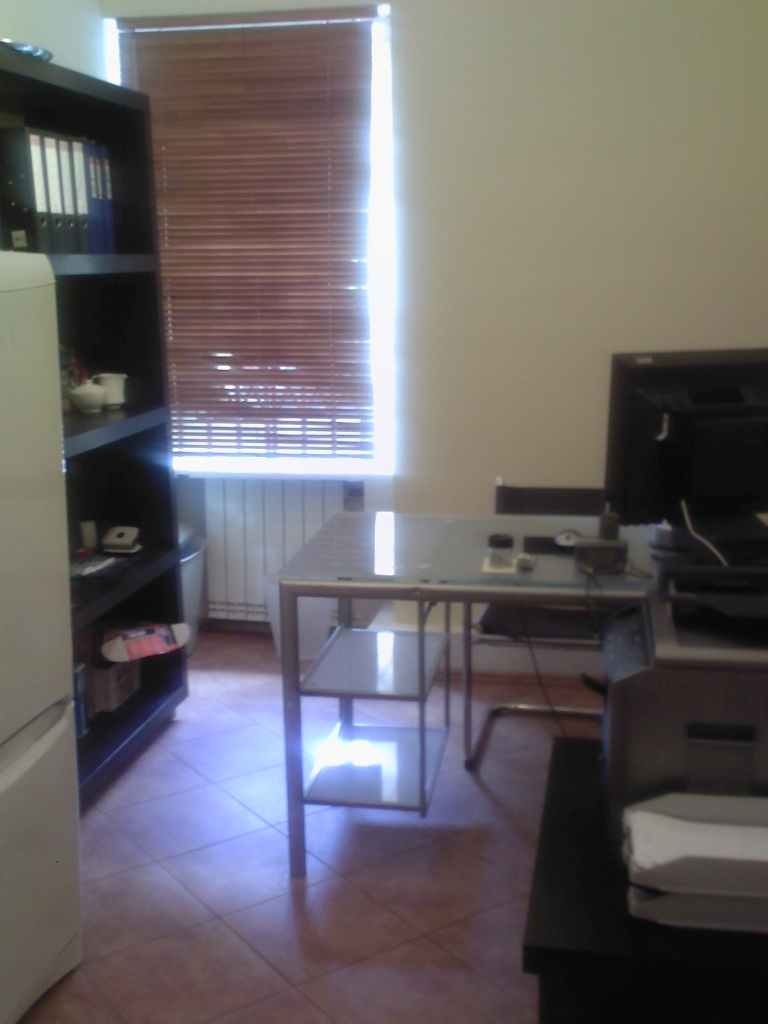 Wnętrze biura z biurkiem, komputerem, regałem z segregatorami i roletami w oknie, widoczne płytki podłogowe.