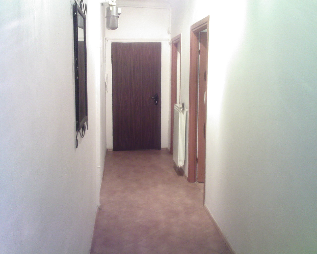 Wnętrze korytarza z brązowymi drzwiami wejściowymi, wiszącym lustrem z metalowymi zdobieniami po lewej stronie, grzejnikiem i otwartymi drzwiami po prawej stronie, oraz brązowymi płytkami na podłodze.