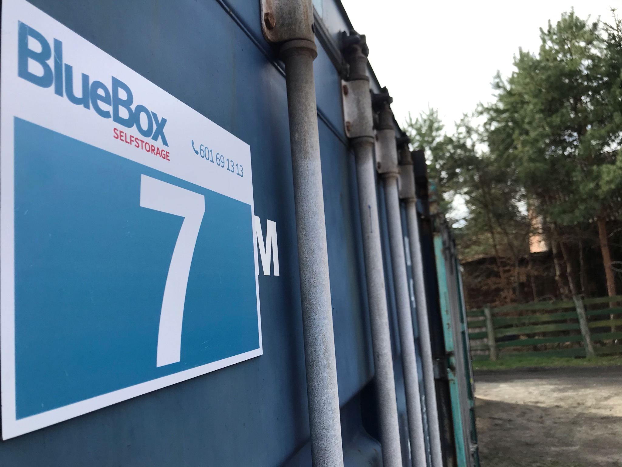 Zewnętrzny kontener magazynowy BlueBox z numerem 7M, widoczne zardzewiałe elementy mocujące, w tle drzewa i ogrodzenie.