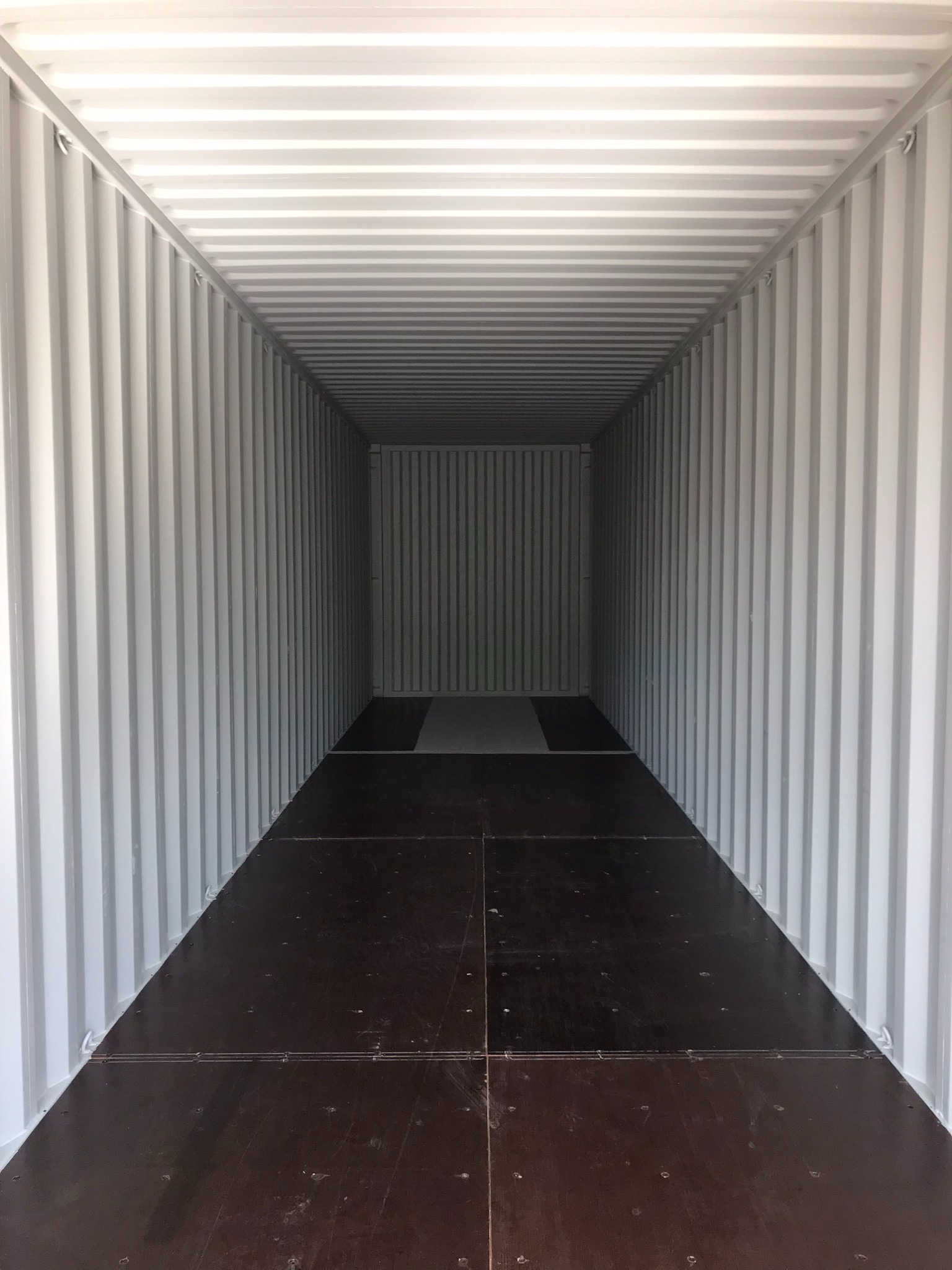 Wnętrze pustego, pomalowanego na biało kontenera transportowego z brązową, drewnianą podłogą. Widok z głębi kontenera w kierunku wyjścia.