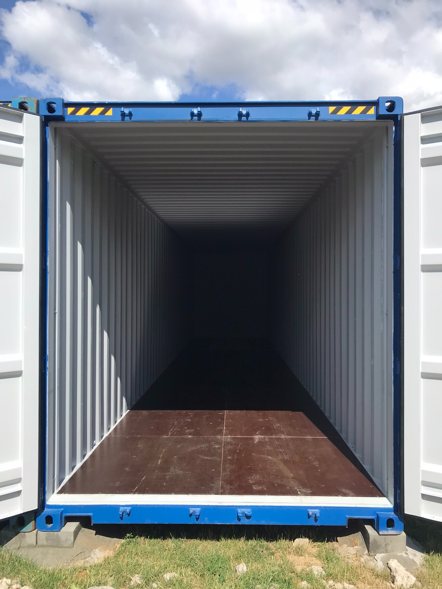 Otwarty, pusty kontener magazynowy z drewnianą podłogą, widok z zewnątrz na wnętrze, błękitne obramowanie, żółto-czarne pasy ostrzegawcze u góry