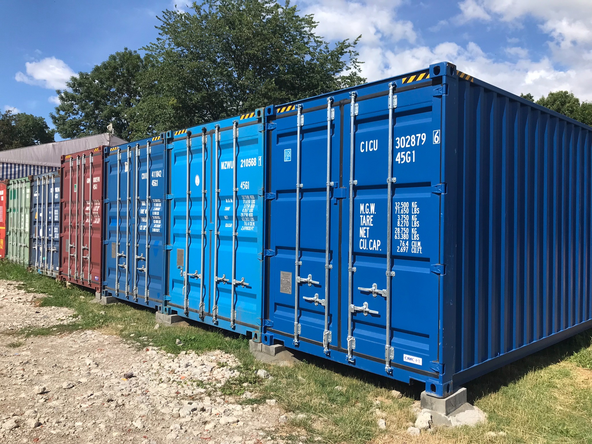 Rząd kolorowych kontenerów magazynowych ustawionych na betonowych podstawach, widoczne oznaczenia i napisy na ścianach, tło stanowią drzewa i błękitne niebo.