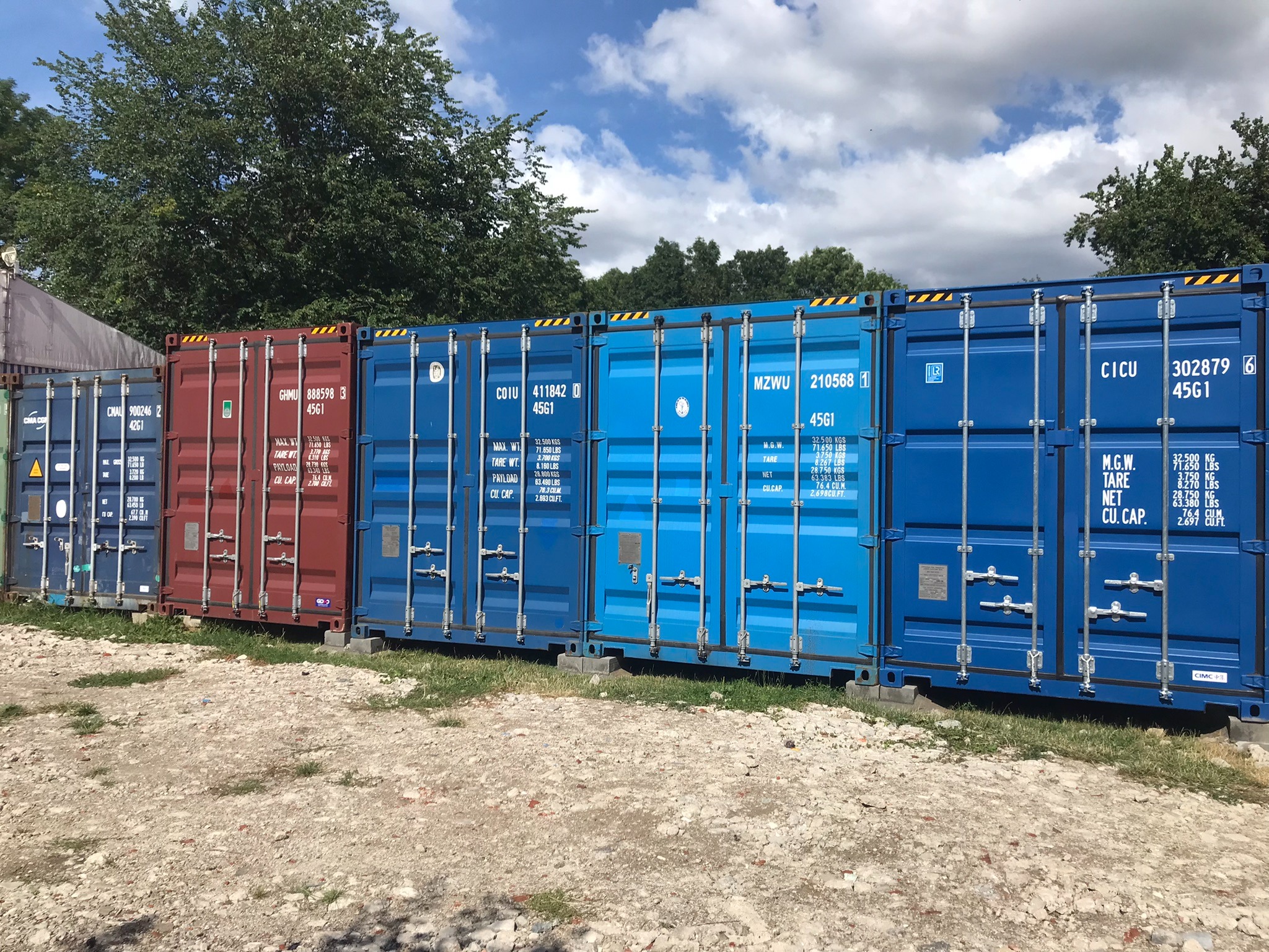 Rząd kolorowych kontenerów magazynowych ustawionych na placu z ziemią i trawą, na tle drzew i błękitnego nieba z chmurami.