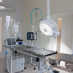 Sala operacyjna z metalowym stołem chirurgicznym, lampą operacyjną, aparaturą do podawania tlenu i kroplówką, przygotowana do zabiegu.