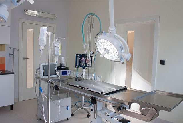 Sala operacyjna z metalowym stołem chirurgicznym, lampą operacyjną, aparaturą do podawania tlenu i kroplówką, przygotowana do zabiegu.