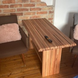 Pracownia Stolarska Podpirka - Stół kawowy z drewna klejonego, z pilotem leżącym na blacie, ustawiony w pokoju z ceglaną ścianą i drewnianą podłogą, obok brązowych foteli z różowymi poduszkami.