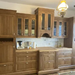 Klasyczna, drewniana zabudowa kuchenna z przeszklonymi szafkami wiszącymi, białymi płytkami na ścianie i ozdobną lampą wiszącą. Na blacie widoczny robot kuchenny i akcesoria.