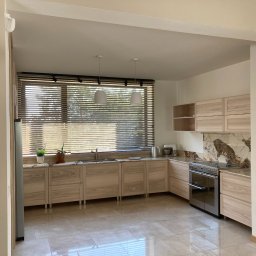 Pracownia Stolarska Podpirka - Jasna kuchnia z drewnianymi frontami, marmurowym blatem i backsplashiem, widok na okno z żaluzjami i dwa białe wiszące światła.