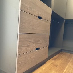 Pracownia Stolarska Podpirka - Szare szafy z drewnianymi frontami szuflad z minimalistycznymi uchwytami, widok na wnętrze garderoby z drążkiem na ubrania, parkiet na podłodze.