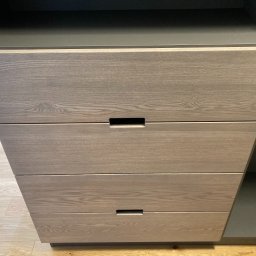 Pracownia Stolarska Podpirka - Szczegółowe ujęcie frontu szafy z czterema szufladami o jasnym, drewnopodobnym wykończeniu i minimalistycznych uchwytach w kształcie podłużnych wgłębień, na tle podłogi z paneli.