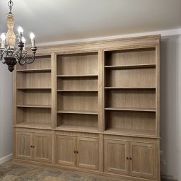 Pracownia Stolarska Podpirka - Duża, drewniana biblioteka na wymiar z szafkami dolnymi i półkami, oświetlona ozdobnym żyrandolem w stylu retro.