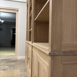 Pracownia Stolarska Podpirka - Duża, drewniana biblioteczka z półkami i szafkami, wykonana z jasnego drewna o wyraźnym usłojeniu, widoczna z boku, z dekoracyjnymi frezowaniami i ciemnymi uchwytami, na tle jasnej ściany i podłogi...