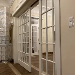 Białe, przeszklone drzwi przesuwne oddzielają pokoje, widoczny parkiet w jodełkę, piec kaflowy zabezpieczony folią ochronną.