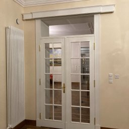 Pracownia Stolarska Podpirka - Białe, przeszklone dwuskrzydłowe drzwi wewnętrzne z ozdobną nadstawką, widoczne z perspektywy korytarza z parkietem w jodełkę, obok grzejnik panelowy i włączniki światła.