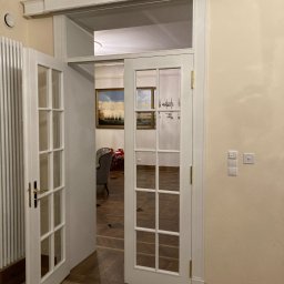 Pracownia Stolarska Podpirka - Białe, przeszklone drzwi wewnętrzne dwuskrzydłowe, z mosiężnymi zawiasami i klamką, częściowo otwarte, widok na parkiet w jodełkę i wnętrze z obrazami i żyrandolem.