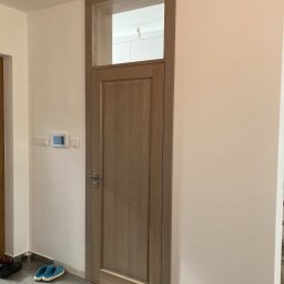 Pracownia Stolarska Podpirka - Zamknięte, drewniane drzwi wewnętrzne z górnym świetlikiem w jasnym korytarzu, widoczne buty i włączniki na ścianie.