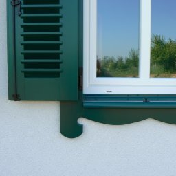 Pracownia Stolarska Podpirka - Białe okno z zielonymi, częściowo otwartymi okiennicami żaluzjowymi, ozdobny parapet zewnętrzny, widok na zieleń za oknem, elewacja o fakturze baranka.