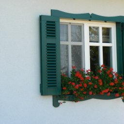 Pracownia Stolarska Podpirka - Białe okno z zielonymi okiennicami i skrzynką z czerwonymi pelargoniami na tle białej ściany.