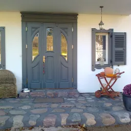 Szare drzwi wejściowe z szybami i dekoracyjnym obramowaniem, uzupełnione o okno z okiennicami, lampę wiszącą i wózek z dyniami na kamiennym chodniku.