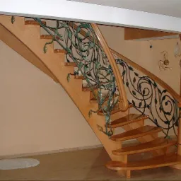 Drewniane schody z ozdobną, metalową balustradą z motywem roślinnym, zakończoną spiralnym elementem, wsparte na kolumnie z podobnym dekorem, w jasnym wnętrzu.