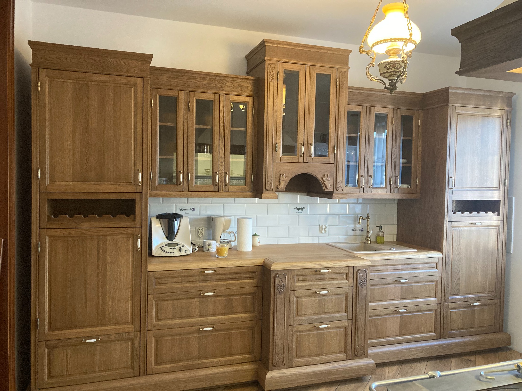 Klasyczna, drewniana zabudowa kuchenna z przeszklonymi szafkami wiszącymi, białymi płytkami na ścianie i ozdobną lampą wiszącą. Na blacie widoczny robot kuchenny i akcesoria.