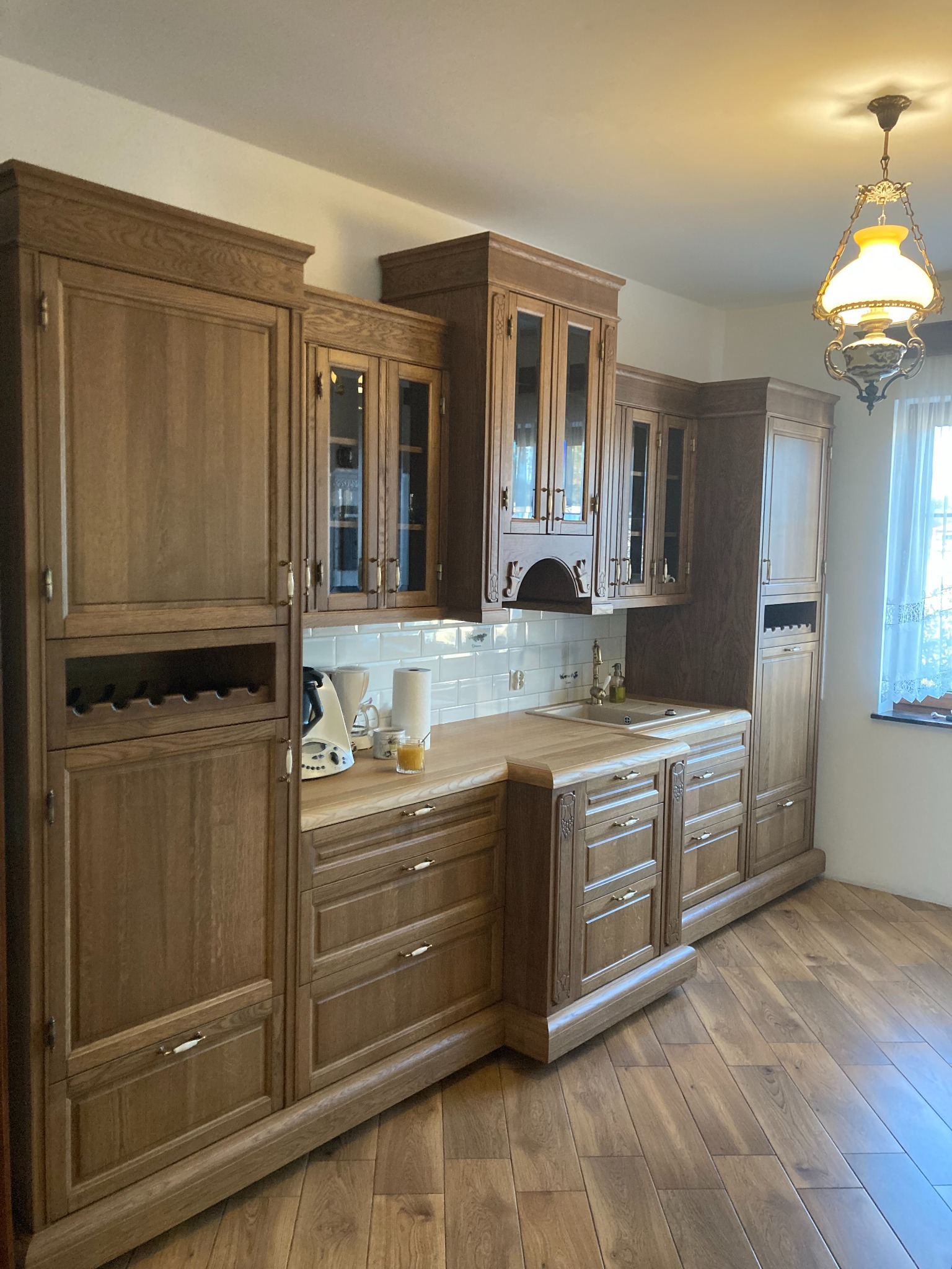 Klasyczna, drewniana zabudowa kuchenna z przeszklonymi szafkami wiszącymi, drewnianym blatem, białymi płytkami na ścianie i wbudowanym regałem na wino, oświetlona ozdobną lampą wiszącą.