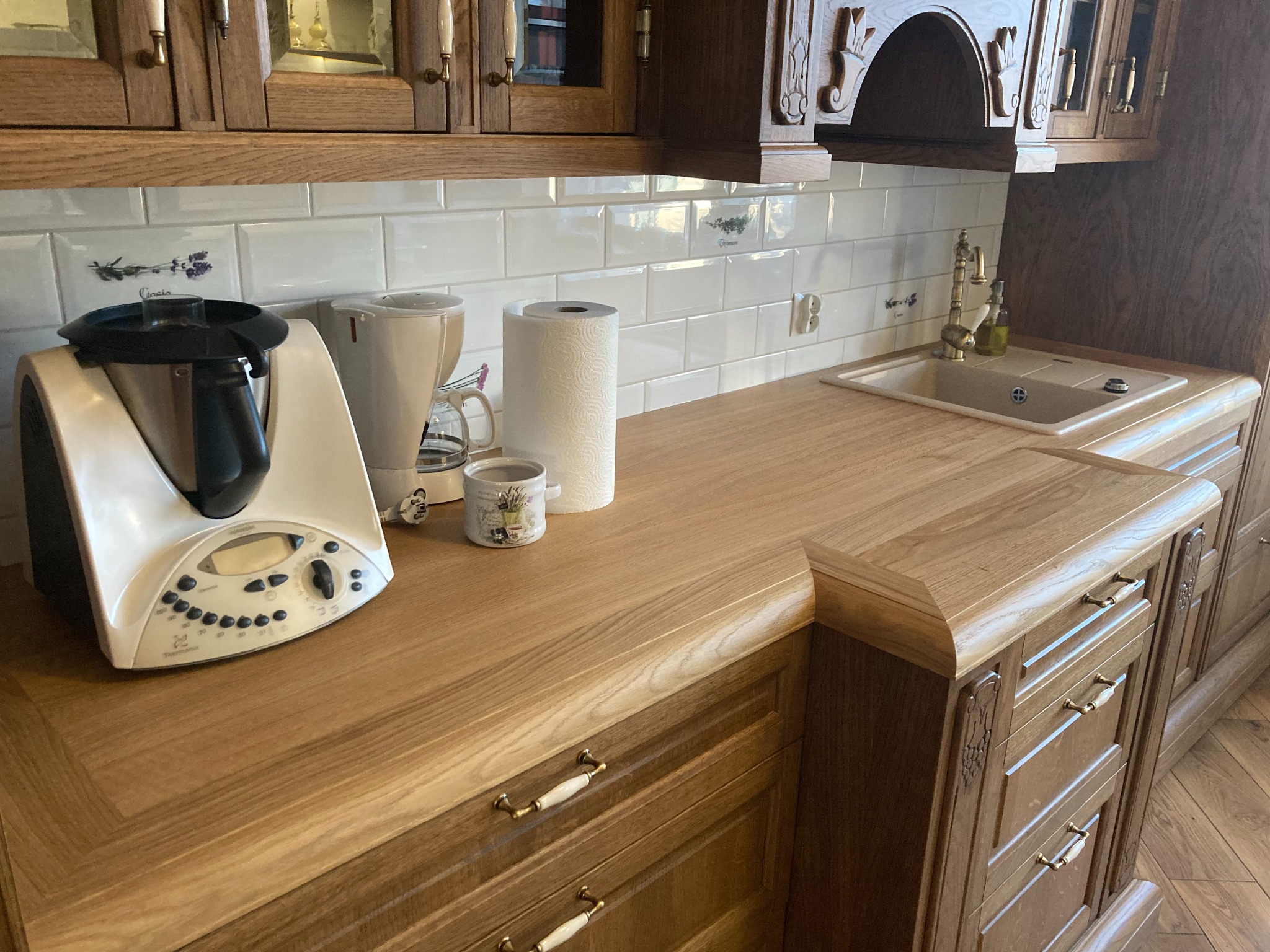 Rustykalna kuchnia z drewnianymi szafkami, blatem z litego drewna, białymi płytkami z motywem lawendy i sprzętami AGD na blacie.