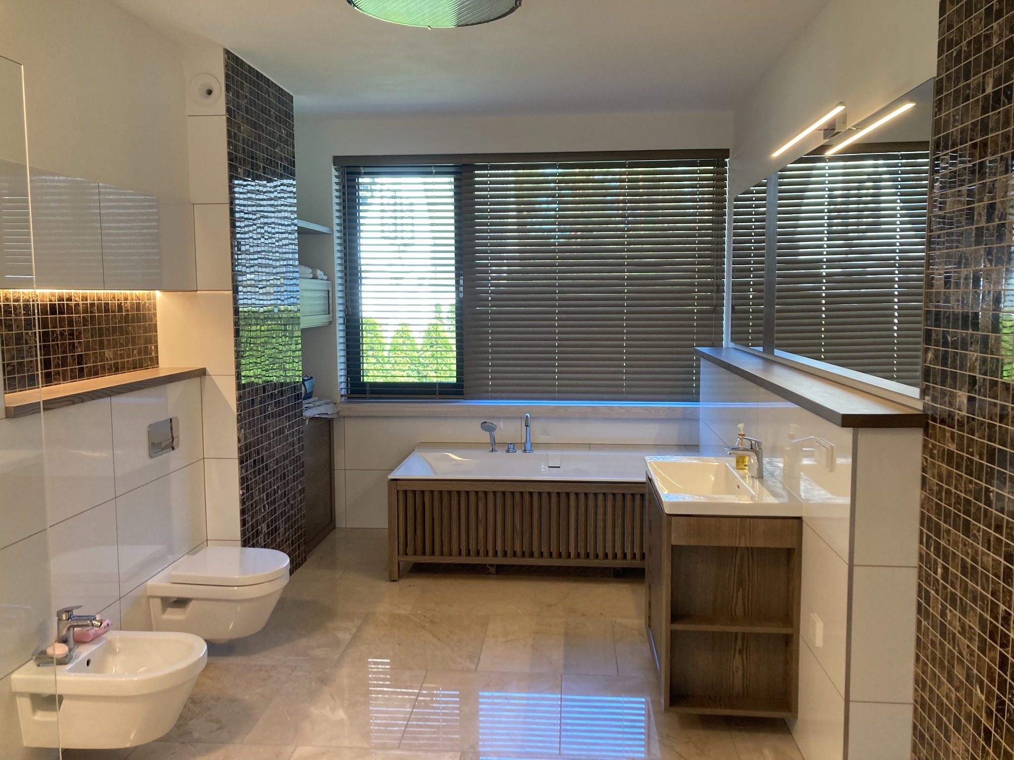 Nowoczesna łazienka z beżowymi płytkami podłogowymi, wiszącą toaletą i bidetem, wanną obudowaną drewnianymi listwami oraz szafką umywalkową z drewnianymi akcentami, oświetlona listwami LED...