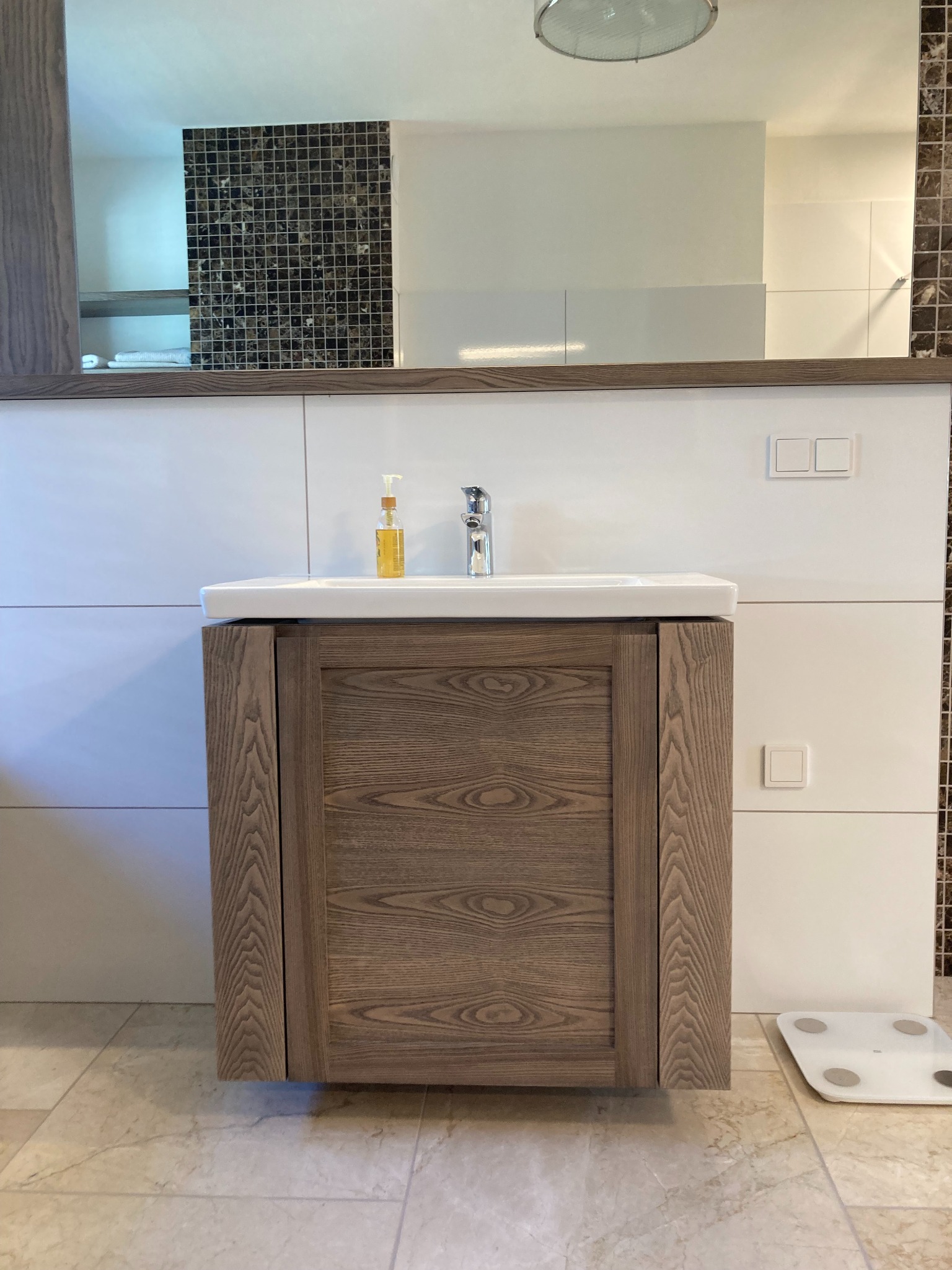 Nowoczesna łazienka z drewnianą szafką pod umywalką, białą ceramiką i mozaiką na ścianie, widoczna waga łazienkowa.