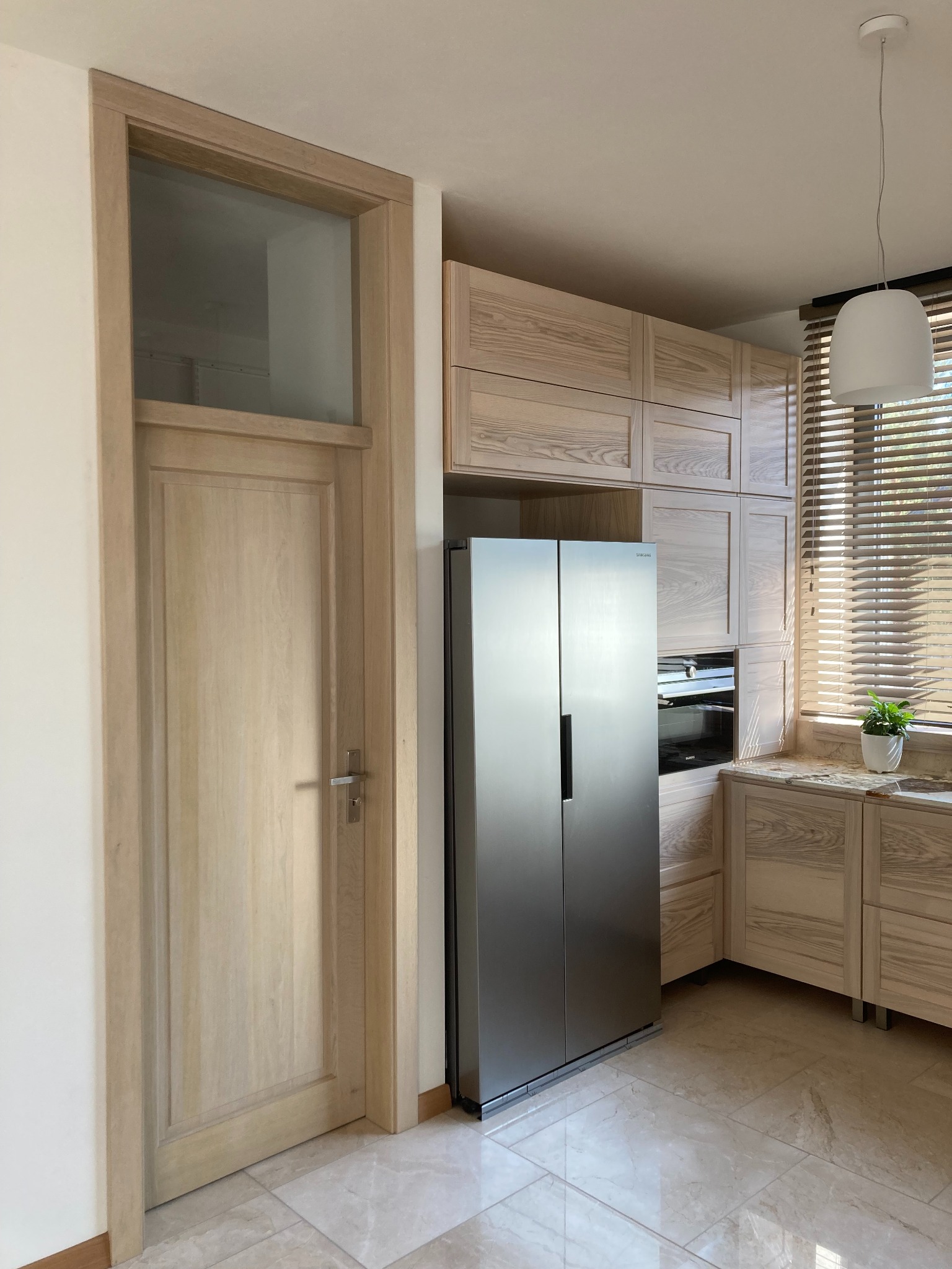 Jasna kuchnia z drewnianymi szafkami, zabudowaną lodówką side-by-side i drzwiami wewnętrznymi z przeszkleniem u góry, podłoga z jasnego marmuru.
