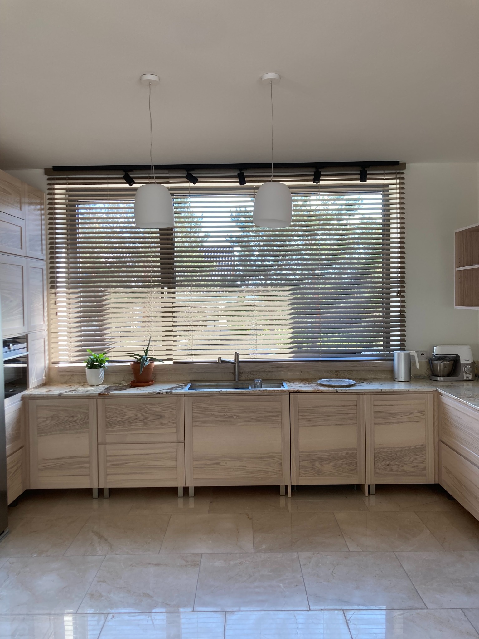Jasna kuchnia z drewnianymi frontami szafek, marmurowym blatem, roletami w oknie i dwoma lampami wiszącymi nad zlewem.