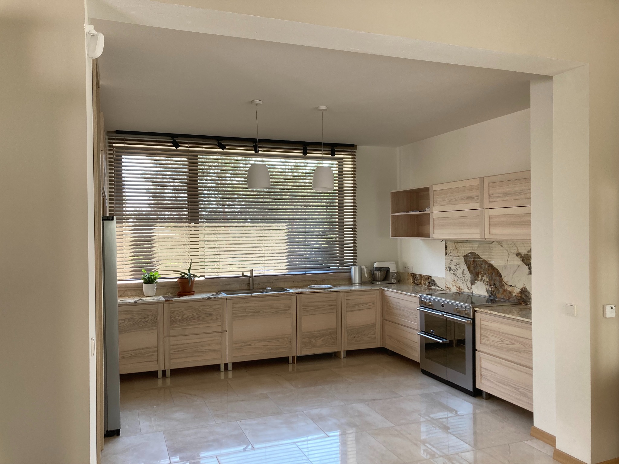 Jasna kuchnia z drewnianymi frontami, marmurowym blatem i backsplashiem, widok na okno z żaluzjami i dwa białe wiszące światła.