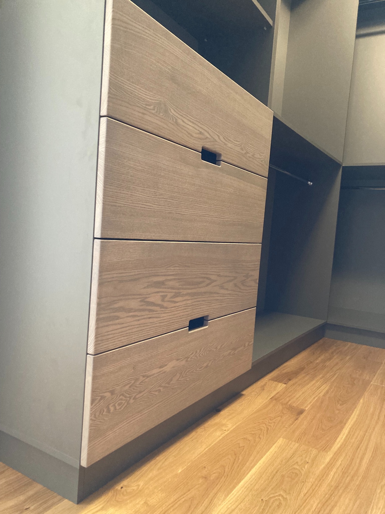 Szare szafy z drewnianymi frontami szuflad z minimalistycznymi uchwytami, widok na wnętrze garderoby z drążkiem na ubrania, parkiet na podłodze.