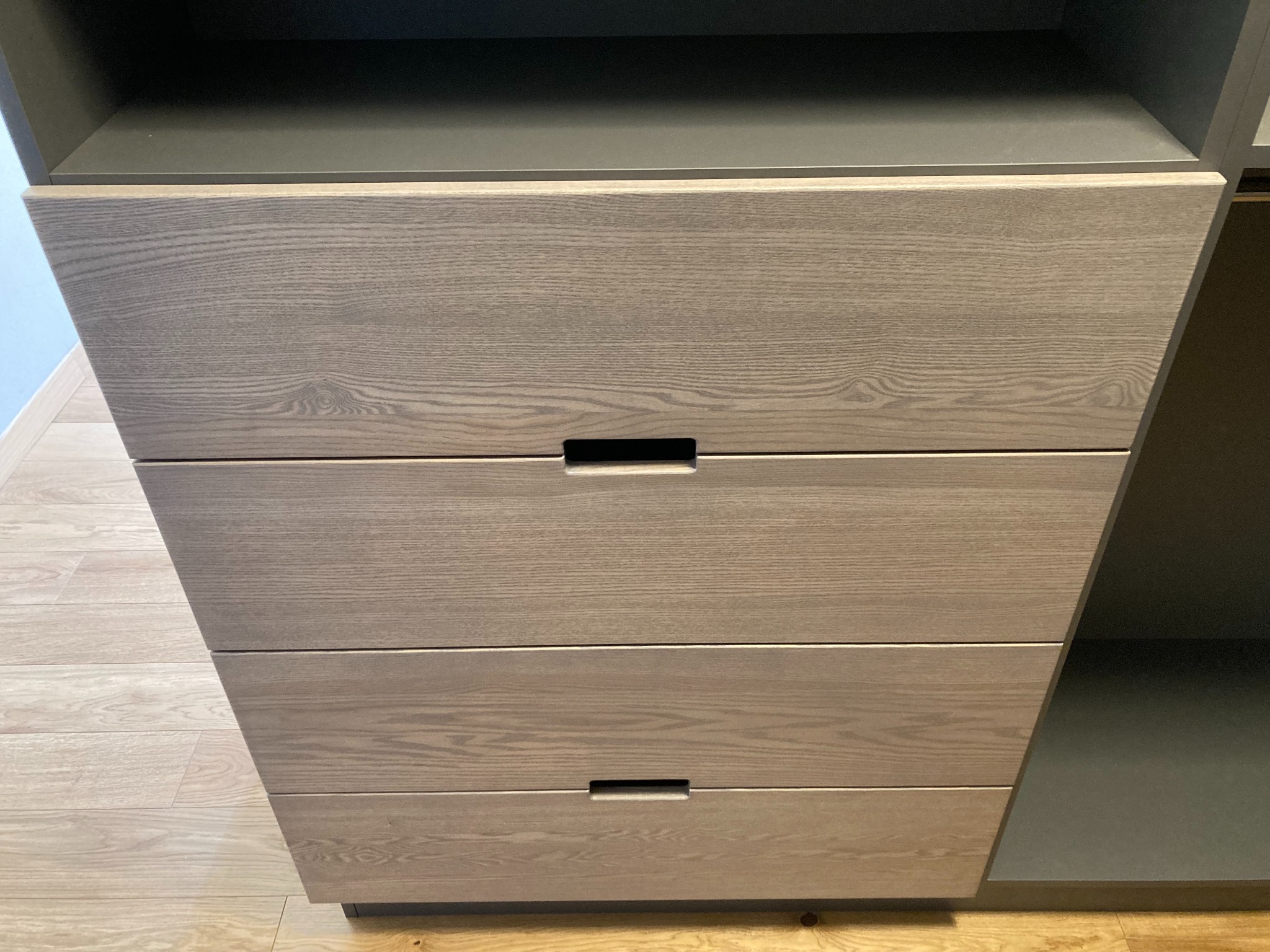 Szczegółowe ujęcie frontu szafy z czterema szufladami o jasnym, drewnopodobnym wykończeniu i minimalistycznych uchwytach w kształcie podłużnych wgłębień, na tle podłogi z paneli.
