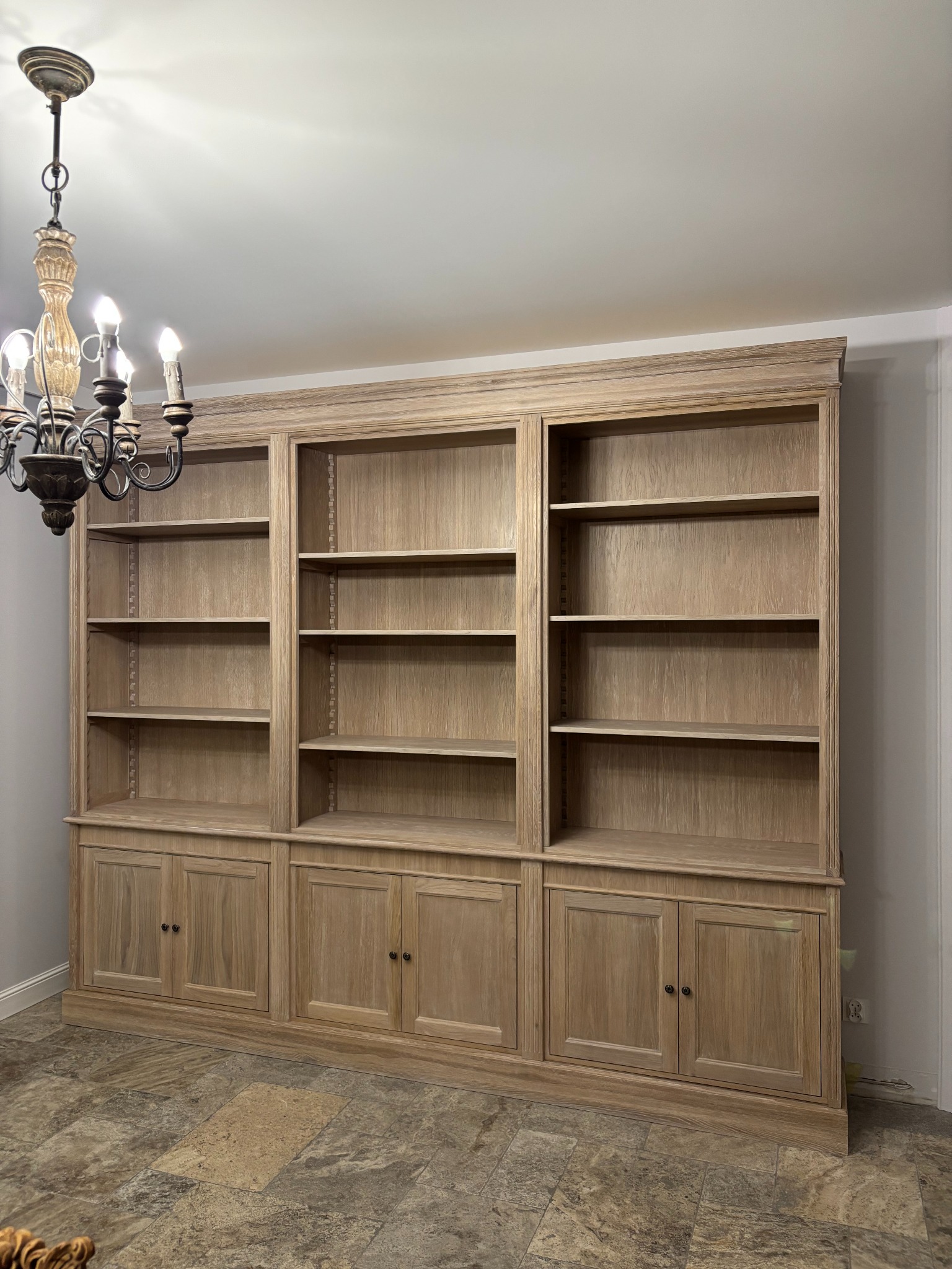 Duża, drewniana biblioteka na wymiar z szafkami dolnymi i półkami, oświetlona ozdobnym żyrandolem w stylu retro.