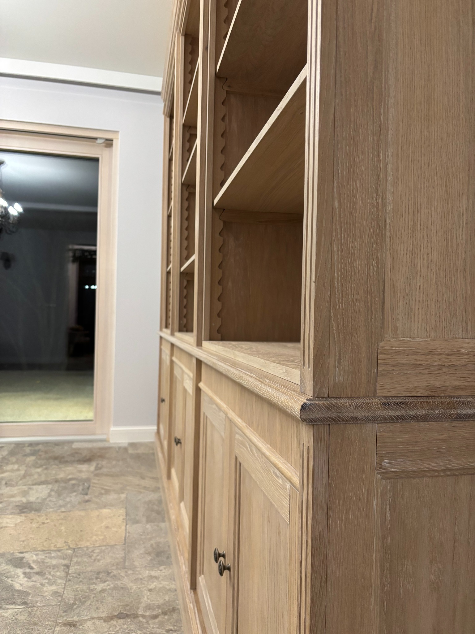 Duża, drewniana biblioteczka z półkami i szafkami, wykonana z jasnego drewna o wyraźnym usłojeniu, widoczna z boku, z dekoracyjnymi frezowaniami i ciemnymi uchwytami, na tle jasnej ściany i podłogi...