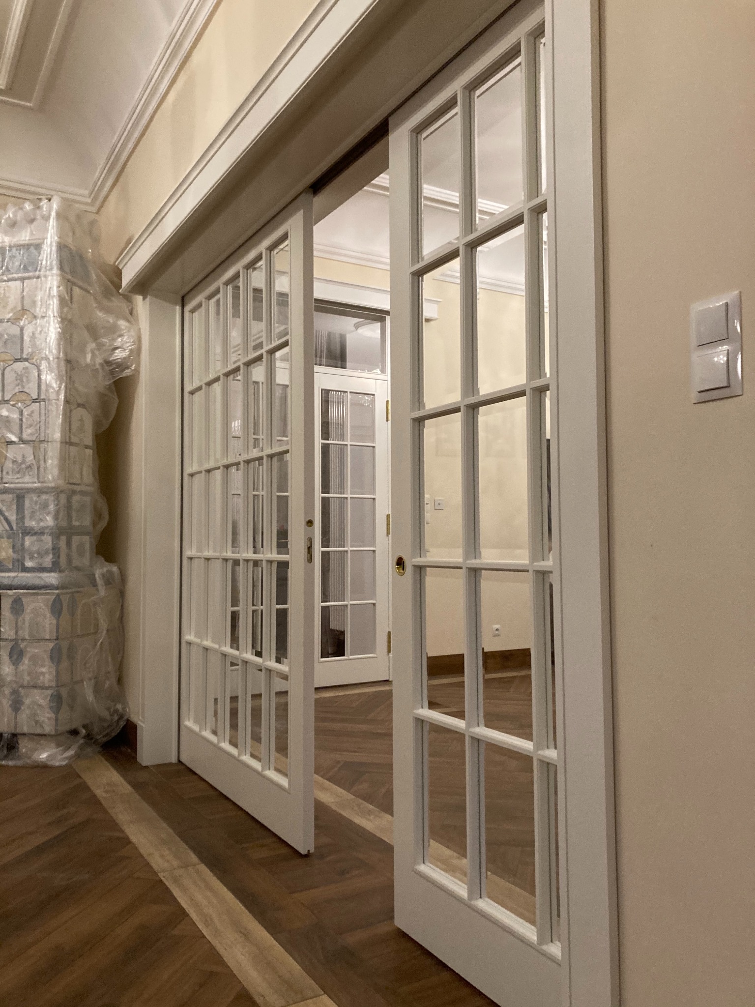 Białe, przeszklone drzwi przesuwne oddzielają pokoje, widoczny parkiet w jodełkę, piec kaflowy zabezpieczony folią ochronną.