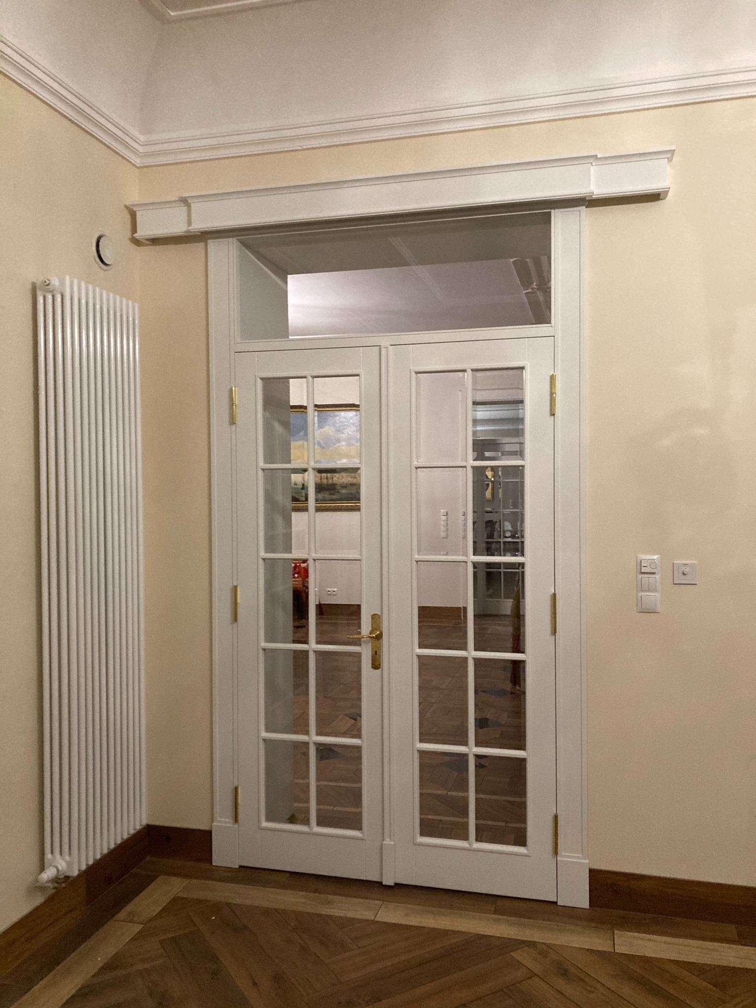 Białe, przeszklone dwuskrzydłowe drzwi wewnętrzne z ozdobną nadstawką, widoczne z perspektywy korytarza z parkietem w jodełkę, obok grzejnik panelowy i włączniki światła.