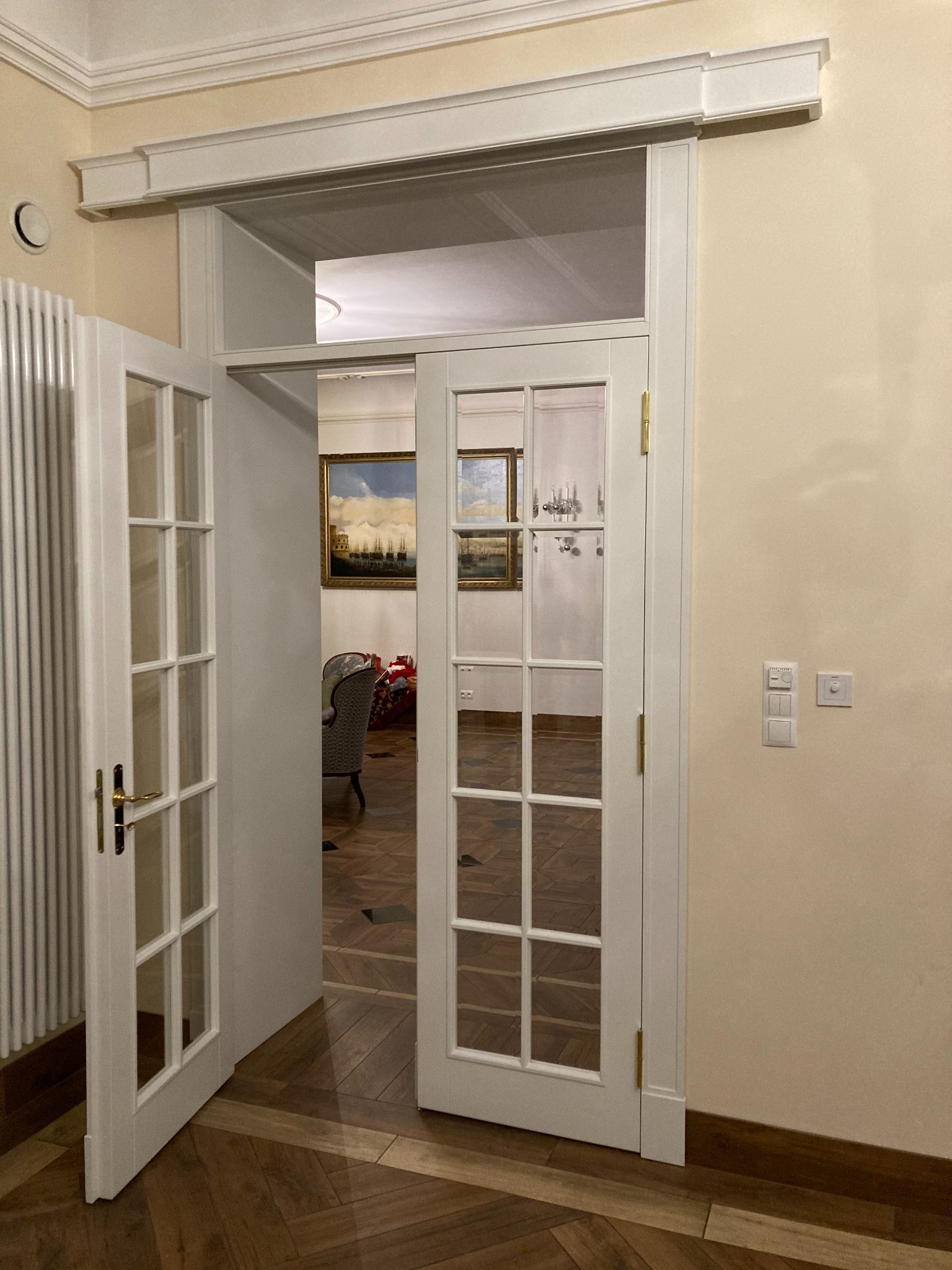 Białe, przeszklone drzwi wewnętrzne dwuskrzydłowe, z mosiężnymi zawiasami i klamką, częściowo otwarte, widok na parkiet w jodełkę i wnętrze z obrazami i żyrandolem.