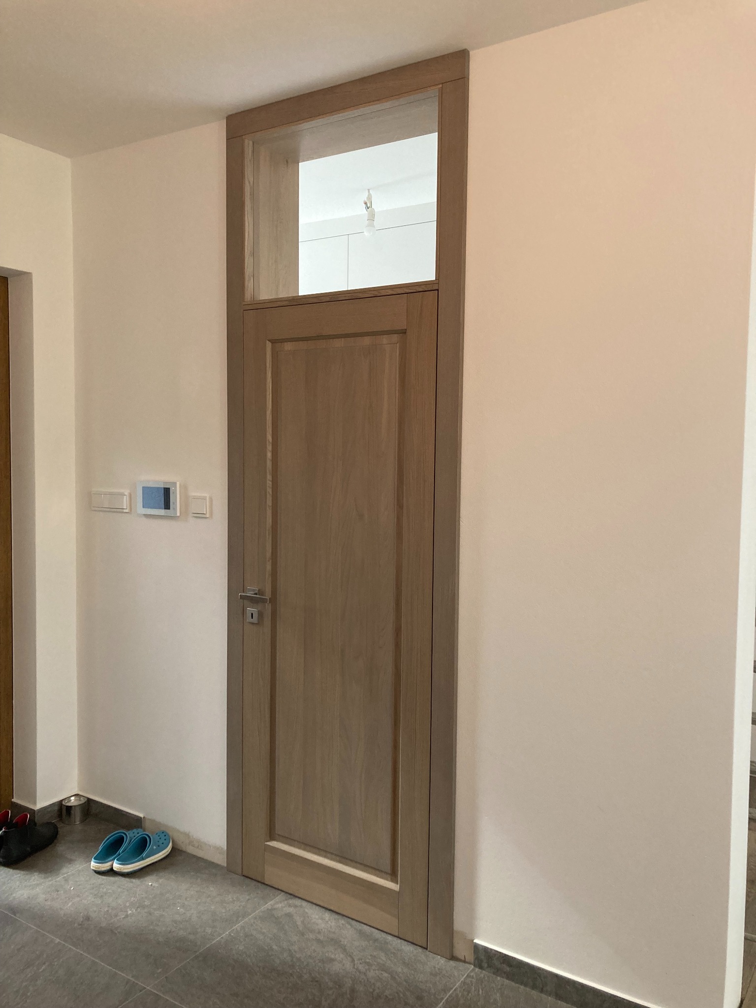 Zamknięte, drewniane drzwi wewnętrzne z górnym świetlikiem w jasnym korytarzu, widoczne buty i włączniki na ścianie.