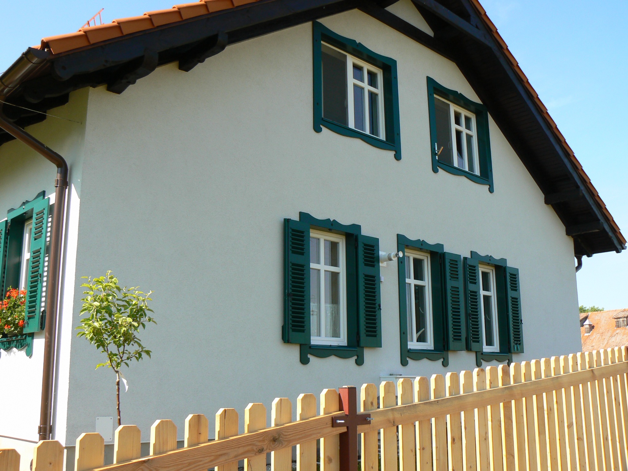 Elewacja domu z białym tynkiem i zielonymi okiennicami, widoczne okna z ozdobnymi ramami i drewniany płot w kolorze naturalnego drewna, małe drzewko przed domem.