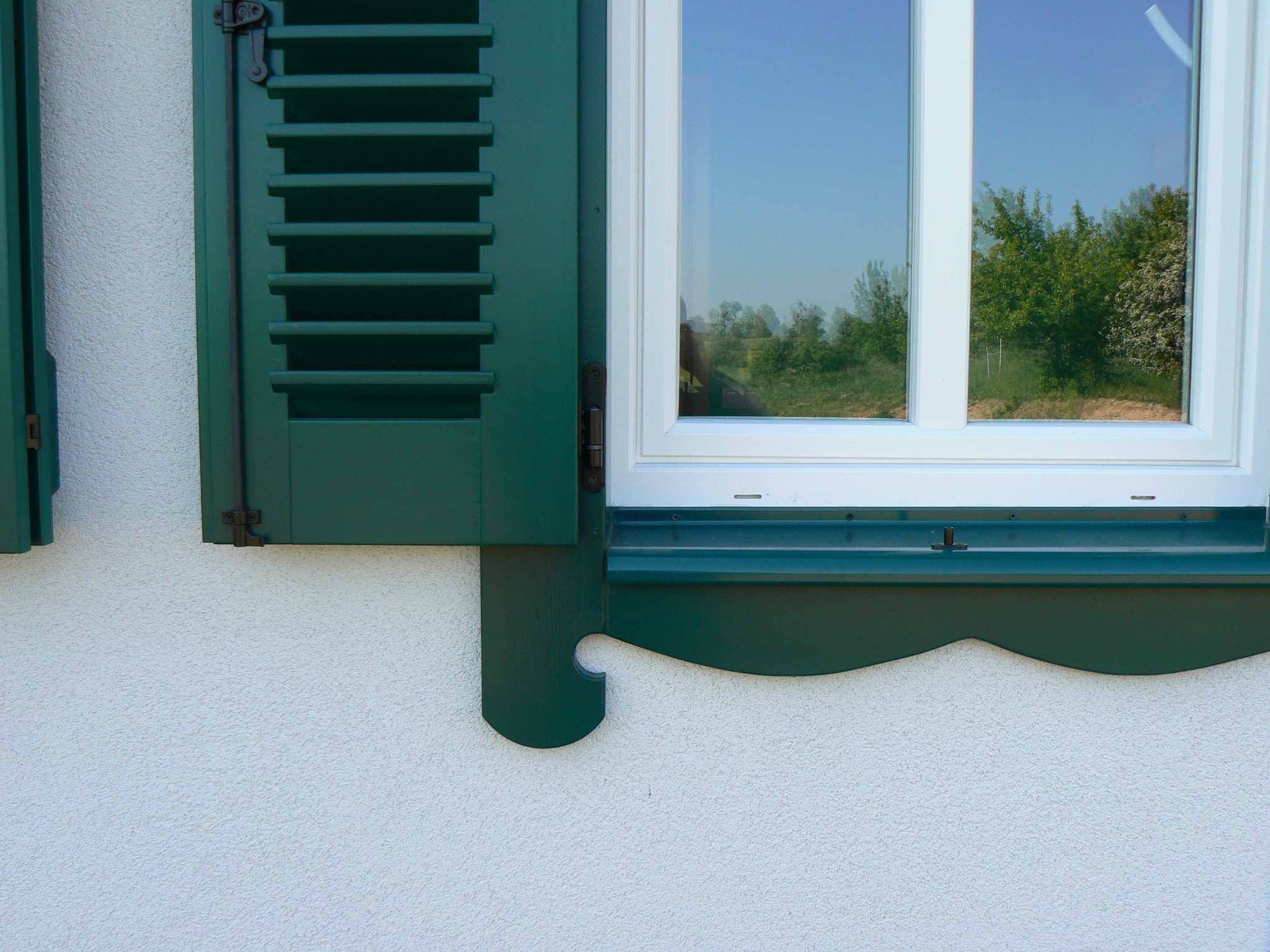 Białe okno z zielonymi, częściowo otwartymi okiennicami żaluzjowymi, ozdobny parapet zewnętrzny, widok na zieleń za oknem, elewacja o fakturze baranka.